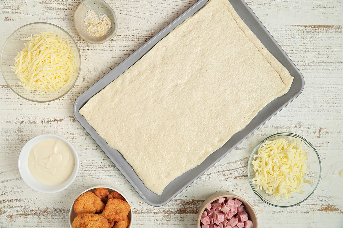A baking tray with rolled-out dough is on a white wooden surface, surrounded by bowls of shredded cheese, cubed ham, breaded chicken pieces, and a creamy white sauce.