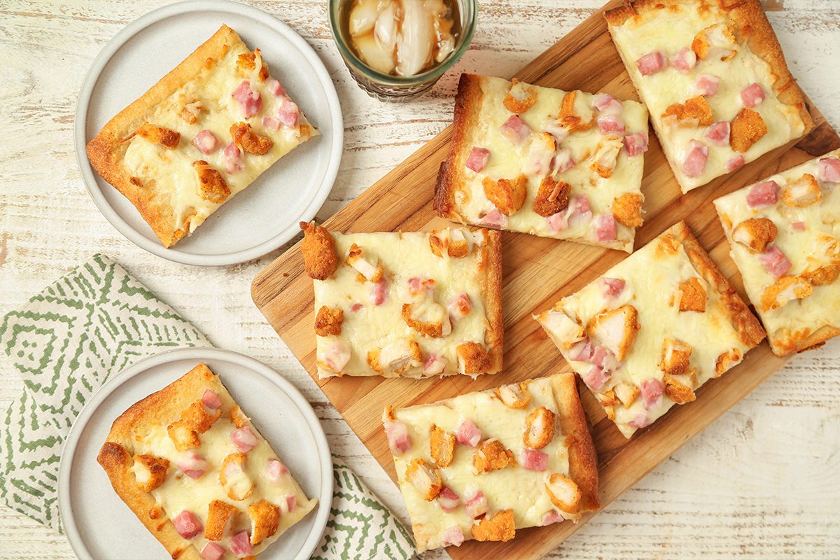 Rectangular slices of baked pizza with cheese, diced ham, and chicken are arranged on a wooden board, with two slices on separate white plates nearby. A patterned napkin and a glass jar are also visible on the table.