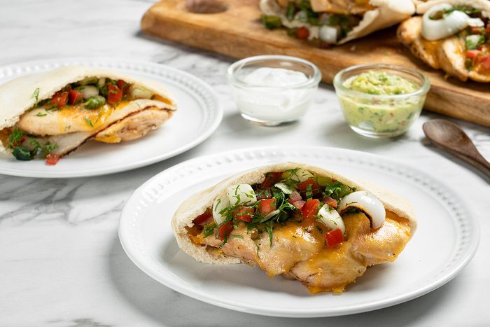 Two plates with grilled chicken breast topped with cheese, onions, tomatoes, and herbs, next to small bowls of sour cream and guacamole on a marble table. A wooden board with more servings is in the background.