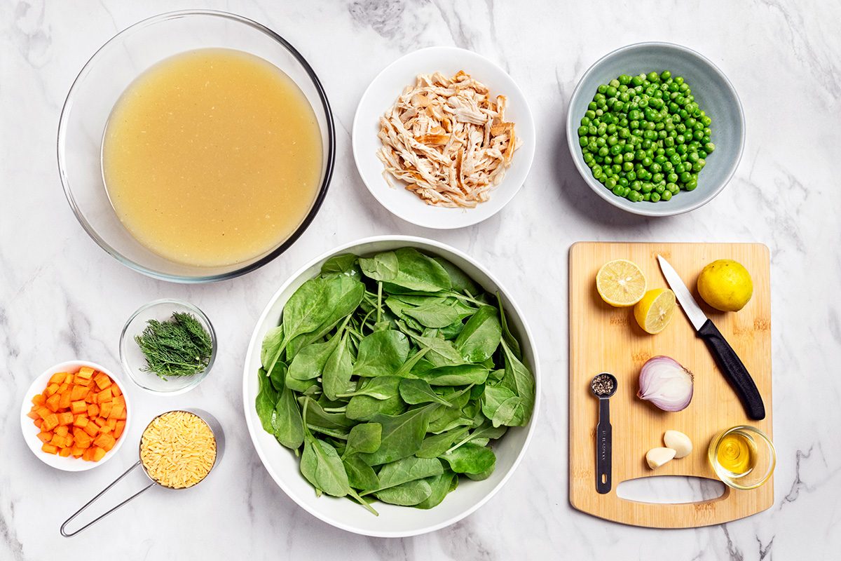 A variety of soup ingredients on a marble surface: a bowl of broth, a bowl of shredded chicken, chopped carrots, fresh spinach, peas, lemon, garlic, shallot, orzo pasta, and fresh herbs on a small cutting board with a knife.