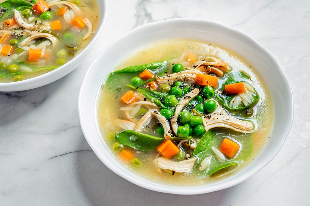 Two bowls of chicken soup with shredded chicken, green peas, carrots, snow peas, and black pepper in a clear broth, set on a white marble surface.