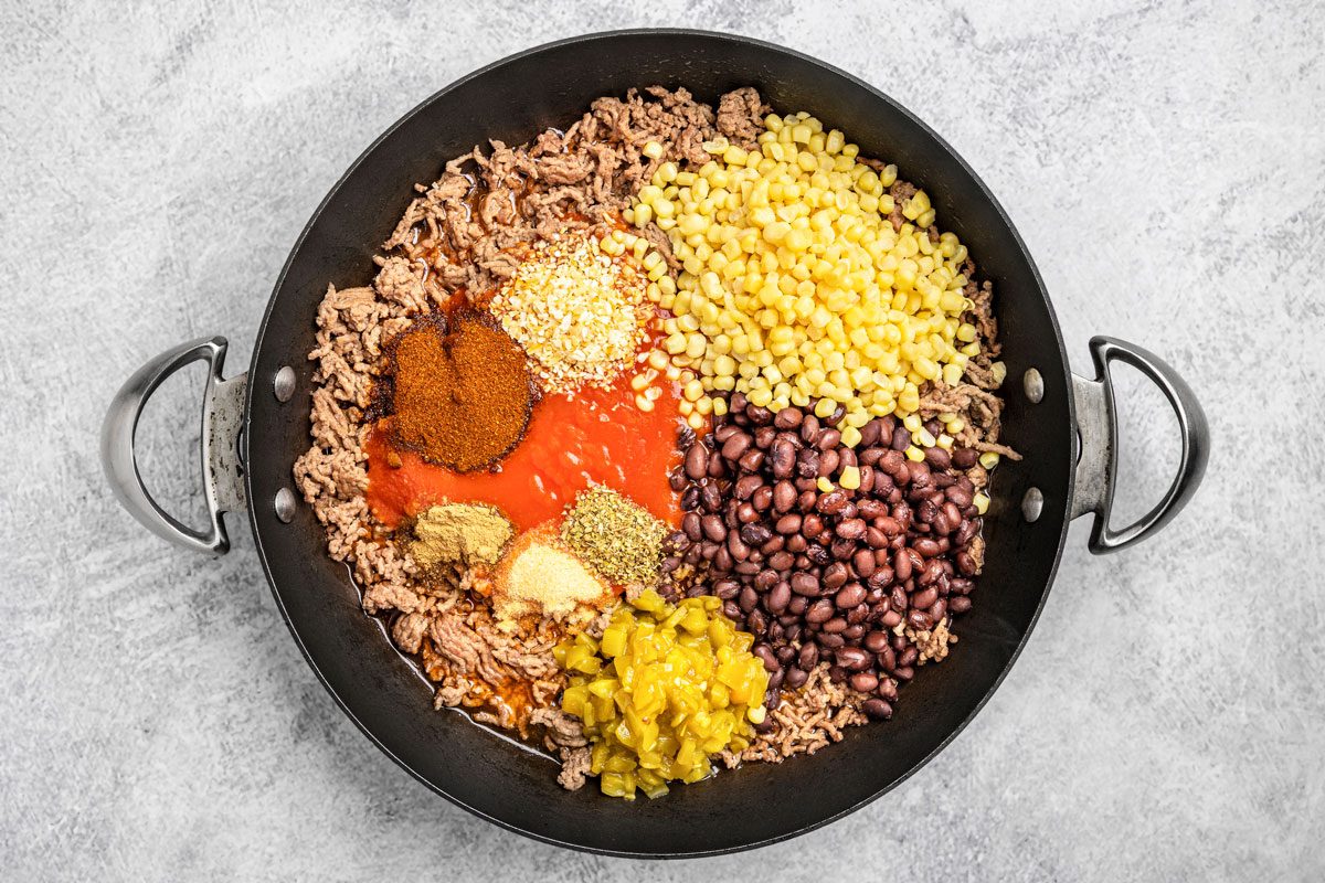 overhead shot of a large black skillet filled with ground beef, black beans, corn, diced green chilies, tomato sauce, and various spices, on a light gray surface