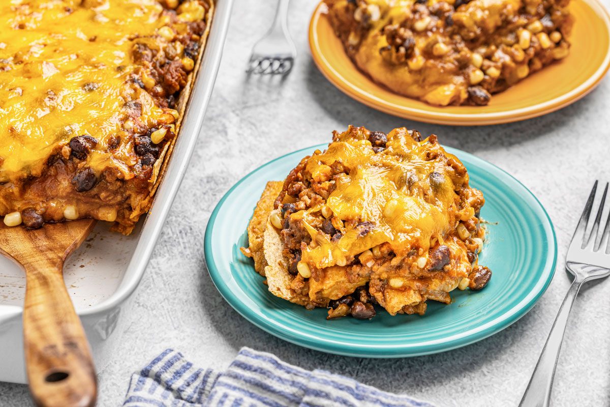 3/4th shot of a slice of chill bake with beans and ground meat is served on a blue plate, with a casserole dish and another serving on an orange plate in the background, along with utensils and a blue striped napkin