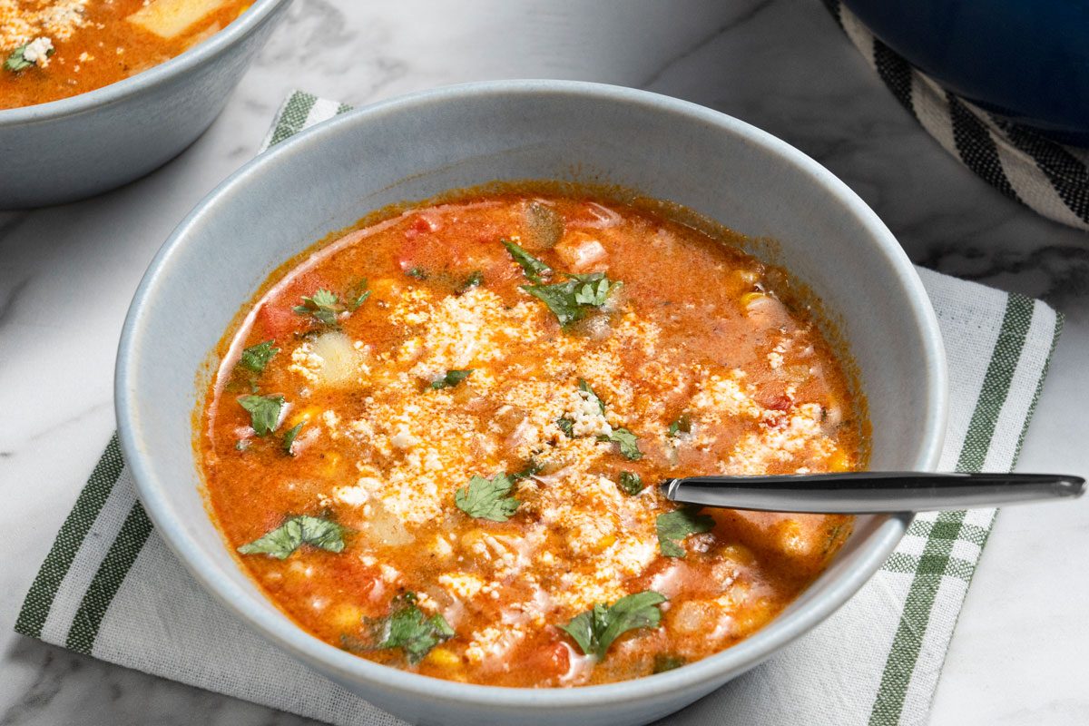 3/4 angle view shot of Mexican Chorizo and Corn Soup served in two light gray bowls; The bowl is placed on a white cloth napkin with green stripes; A spoon rests inside the bowl; all set on a marble surface;