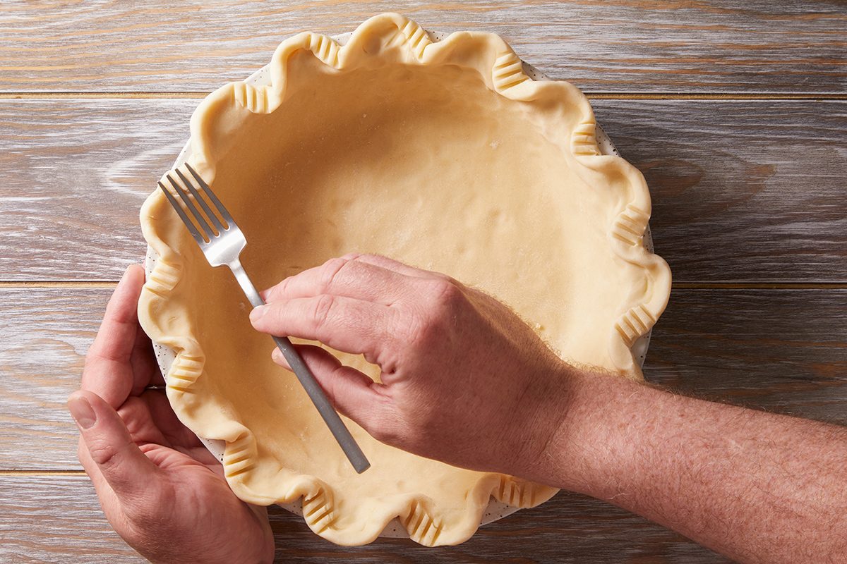 A person uses a fork to crimp the edge of an unbaked pie crust in a pie dish on a wooden surface.