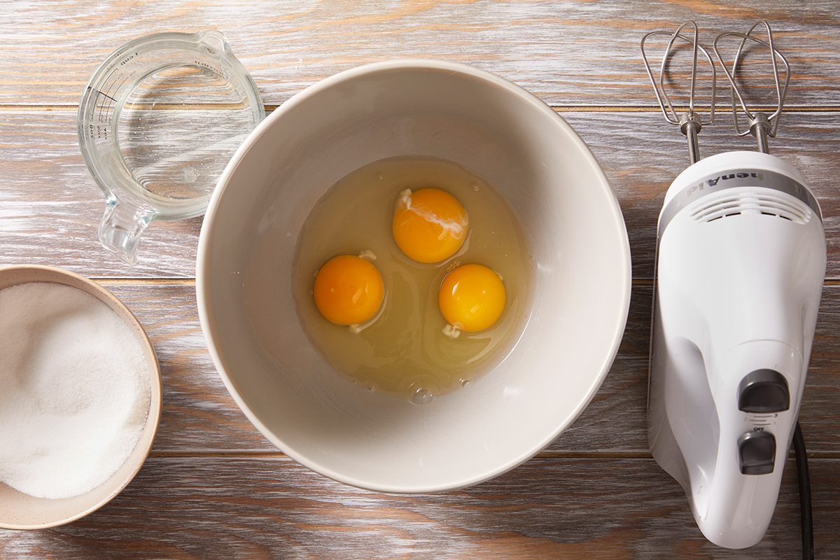 A mixing bowl with three cracked eggs, a bowl of sugar, a measuring cup with liquid, and a hand mixer on a wooden surface.