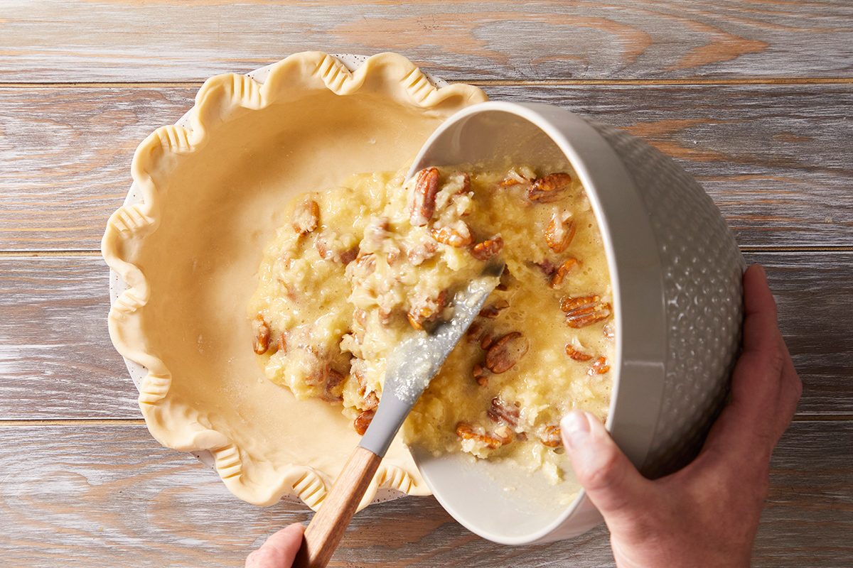 A person spooning a mixture of chopped nuts and filling from a bowl into an unbaked pie crust on a wooden surface.