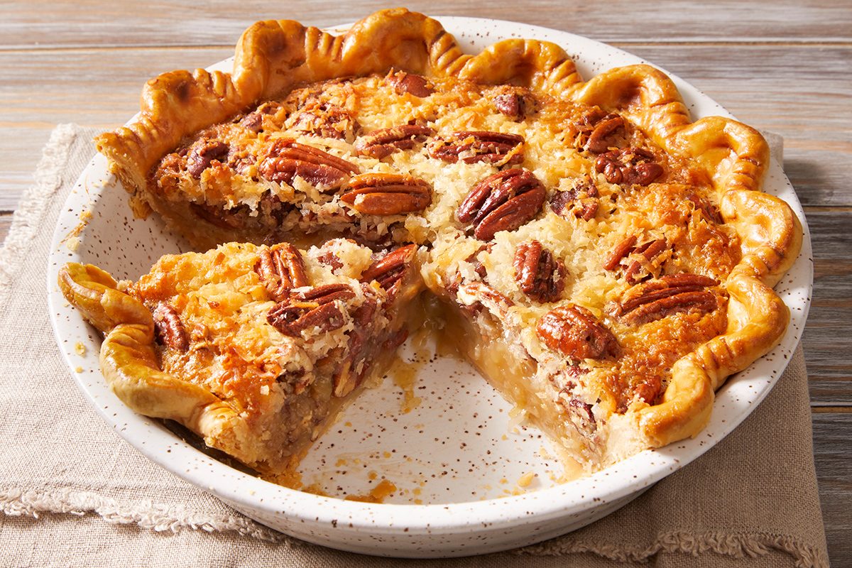 A baked pecan pie in a ceramic pie dish with a golden, flaky crust. A slice has been removed, revealing the gooey, nut-filled filling. The pie rests on a cloth napkin atop a wooden surface.