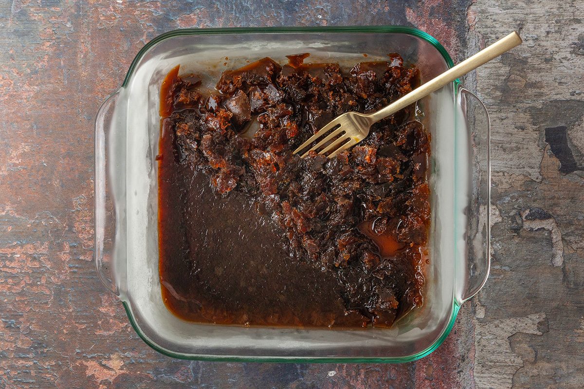 overhead shot of a glass baking dish containing dark brown, partially mashed mixture with a fork resting inside; The mixture appears thick and sticky, covering about half the dish; The background is a rustic surface