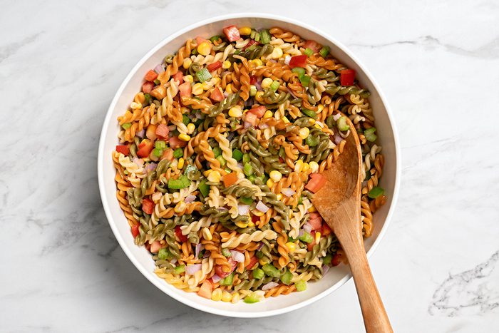A bowl of colorful pasta salad with chopped vegetables, including tomatoes and green peppers, is shown on a marble surface. A wooden spoon rests inside the bowl.