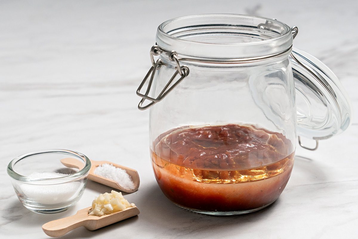 A glass jar with a hinged lid containing a reddish liquid sits on a white surface. Nearby are a small bowl of sugar, a wooden spoon with salt, and another spoon with minced garlic.