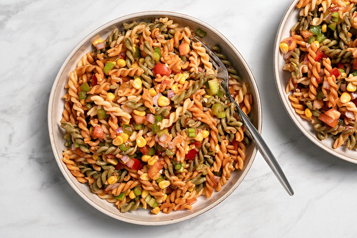 A bowl of tri-color rotini pasta salad mixed with diced tomatoes, corn, and green peppers sits on a marble surface, with a fork resting in the bowl.