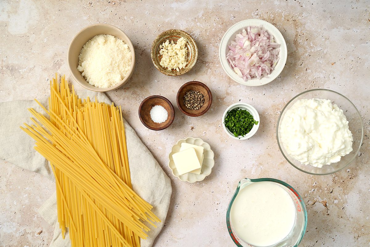Uncooked spaghetti, grated cheese, minced garlic, chopped shallots, salt, pepper, chopped herbs, butter, ricotta cheese, and cream are arranged in small bowls on a beige countertop.