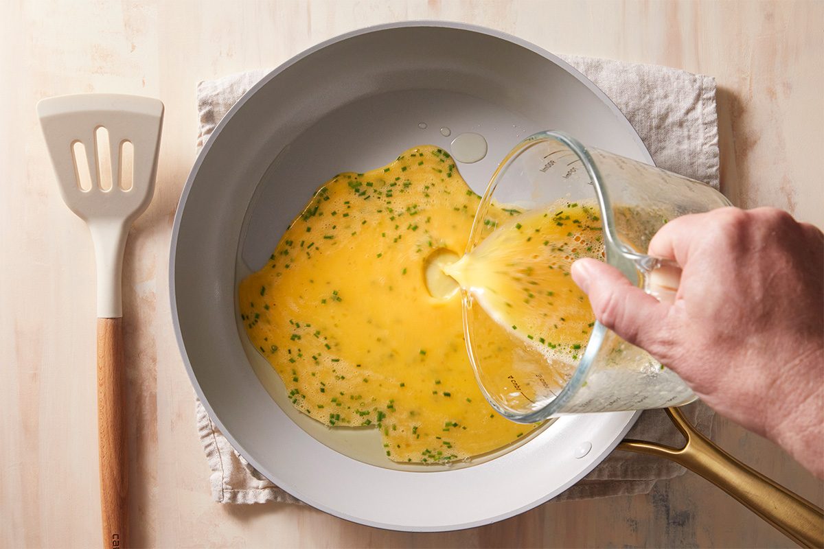 A hand pours beaten eggs with herbs from a glass measuring cup into a white frying pan on a stovetop; a spatula rests nearby on a folded cloth.