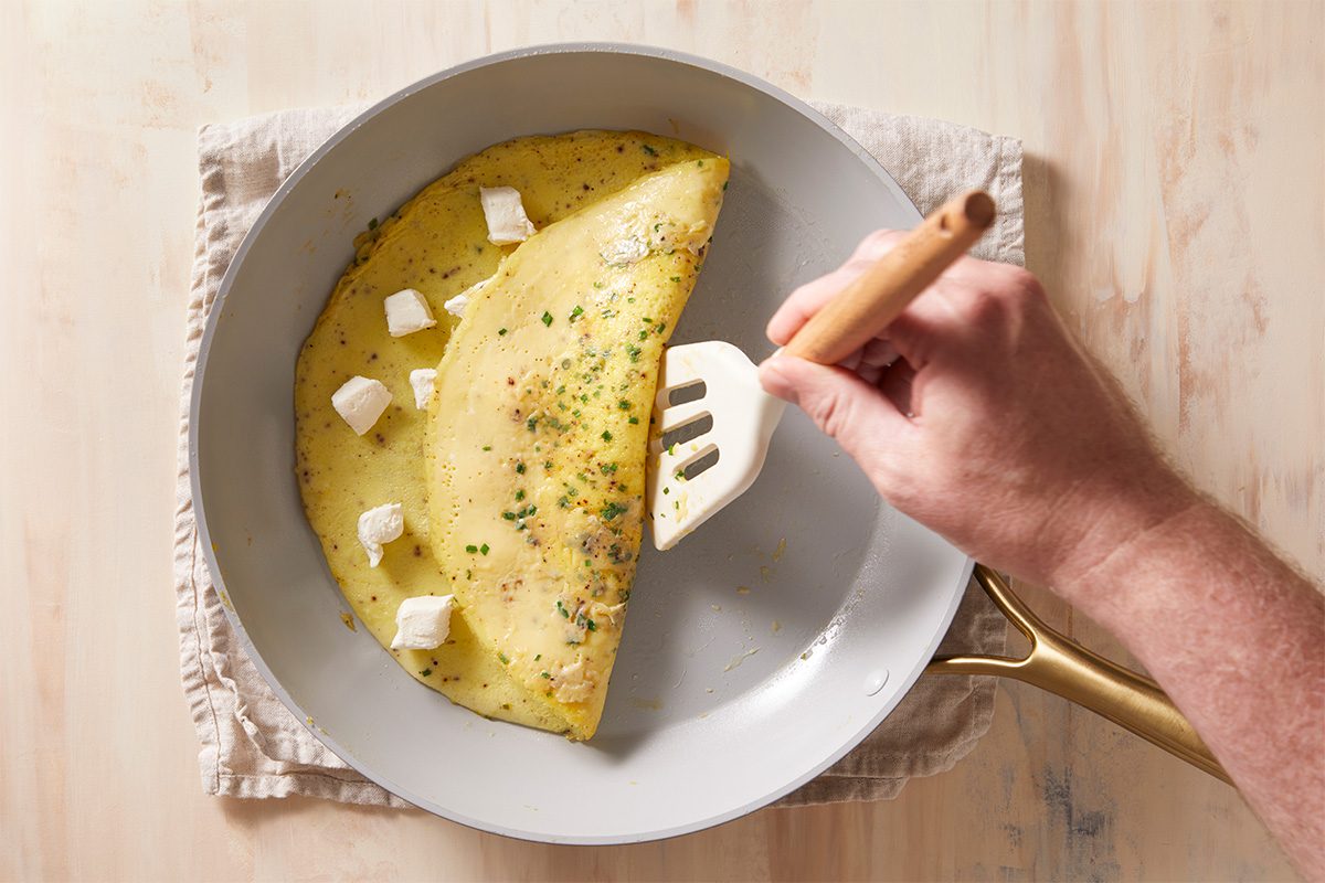 A hand uses a spatula to fold a yellow omelette in a frying pan, with small cubes of white cheese and chopped herbs visible inside, on a beige cloth on a wooden surface.