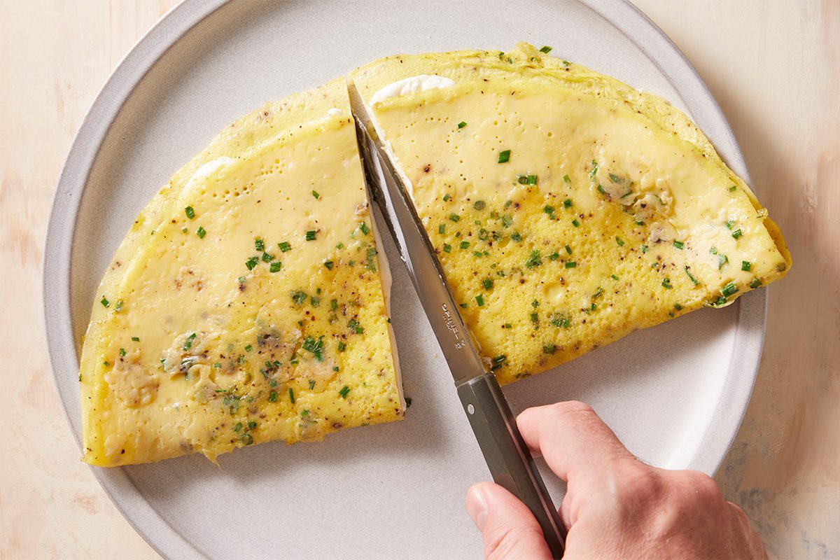 A hand uses a knife to cut a folded omelette with herbs on a white plate.