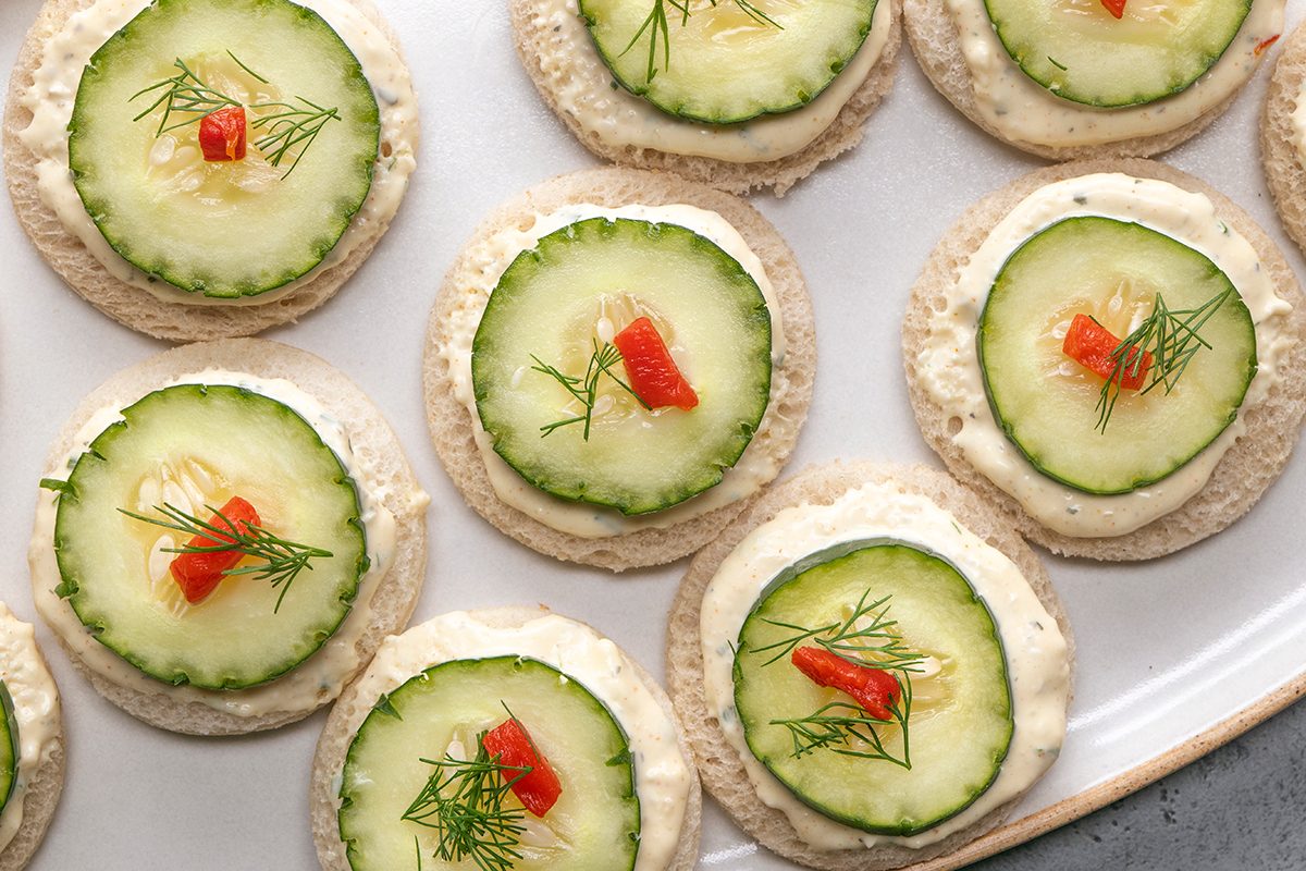 Round crackers topped with a spread, cucumber slices, small pieces of red pepper, and fresh dill are neatly arranged on a white plate.