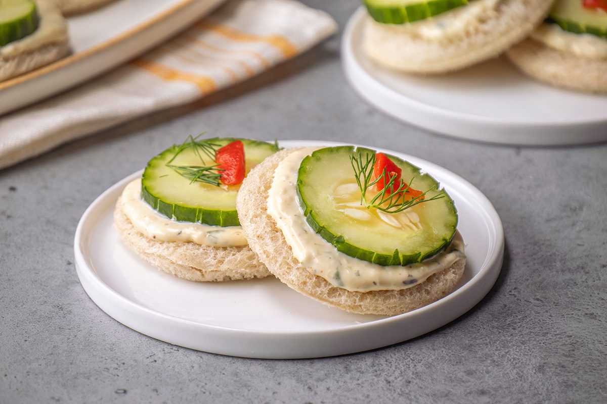 Two round tea sandwiches topped with cream cheese, cucumber slices, a small piece of red pepper, and dill, served on a white plate.