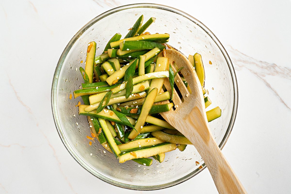 A glass bowl filled with thinly sliced cucumber sticks mixed with seasonings, next to a wooden serving fork and spoon, on a white marble surface.