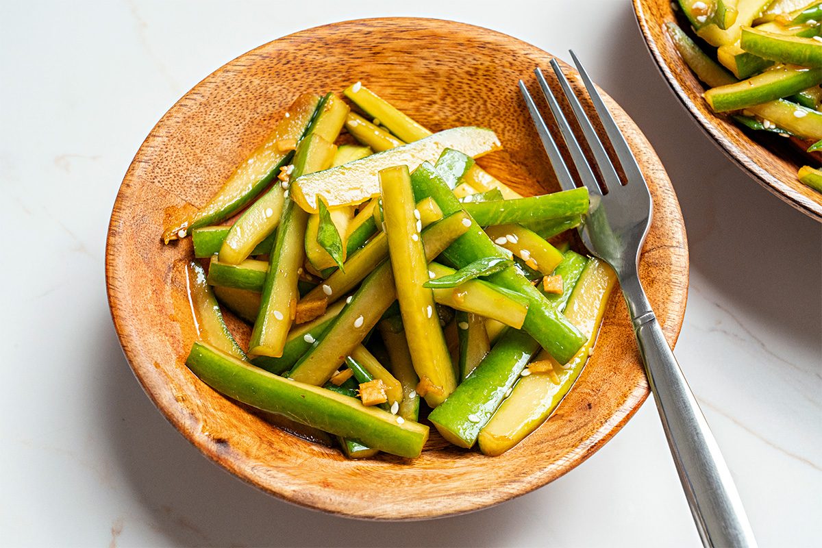 A wooden bowl filled with sliced cucumber salad, garnished with sesame seeds and a light dressing. A fork rests on the edge of the bowl, all placed on a white surface.