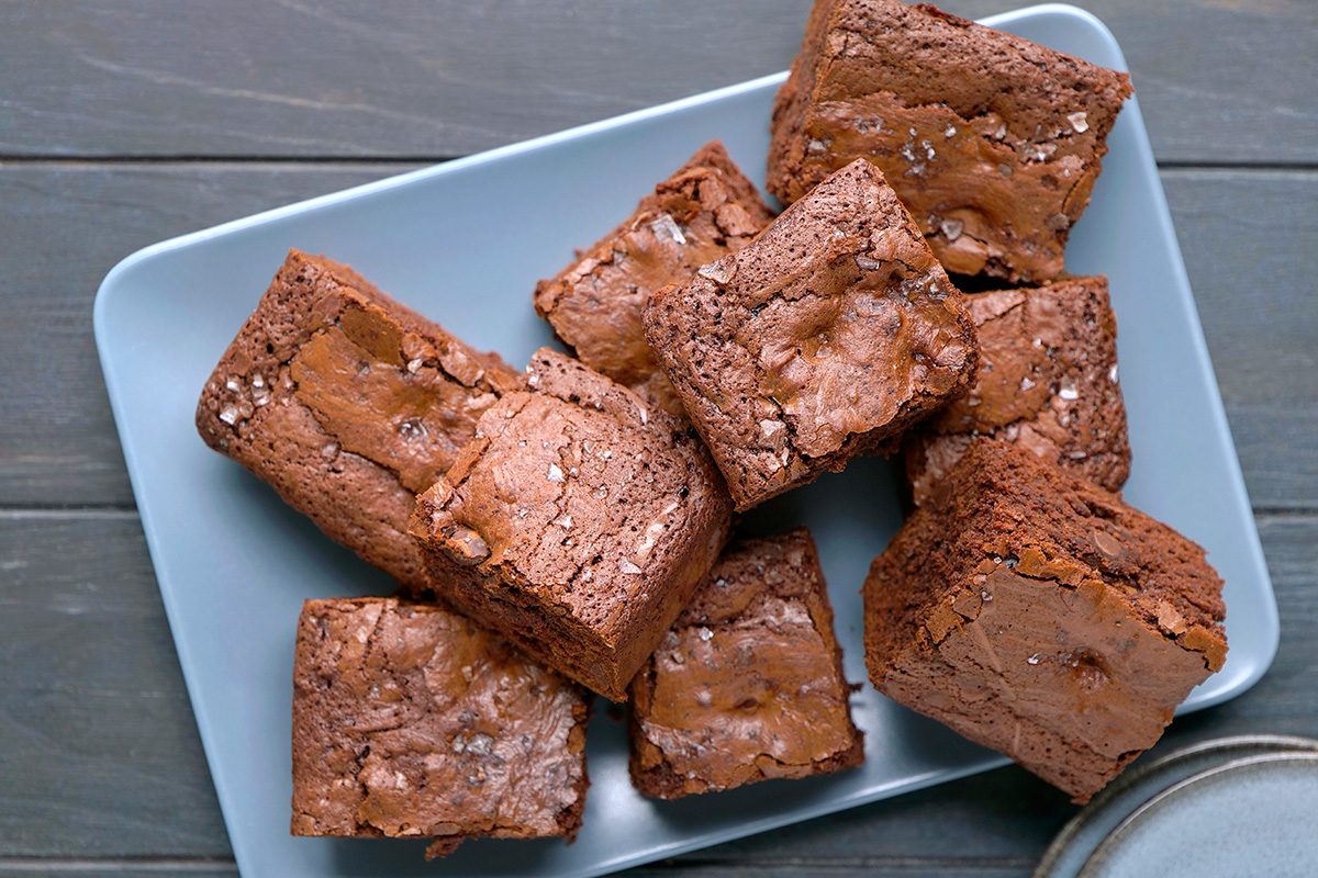 A rectangular blue plate with several thick, fudgy chocolate brownies stacked and overlapping, topped with a light sprinkle of sea salt, on a dark wooden surface.