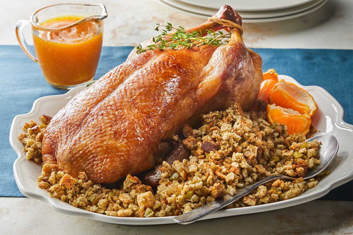 A roasted duck sits on a platter atop a bed of stuffing, garnished with fresh herbs and orange slices. A spoon rests on the side, and a small glass pitcher of sauce is nearby. Plates are stacked in the background.