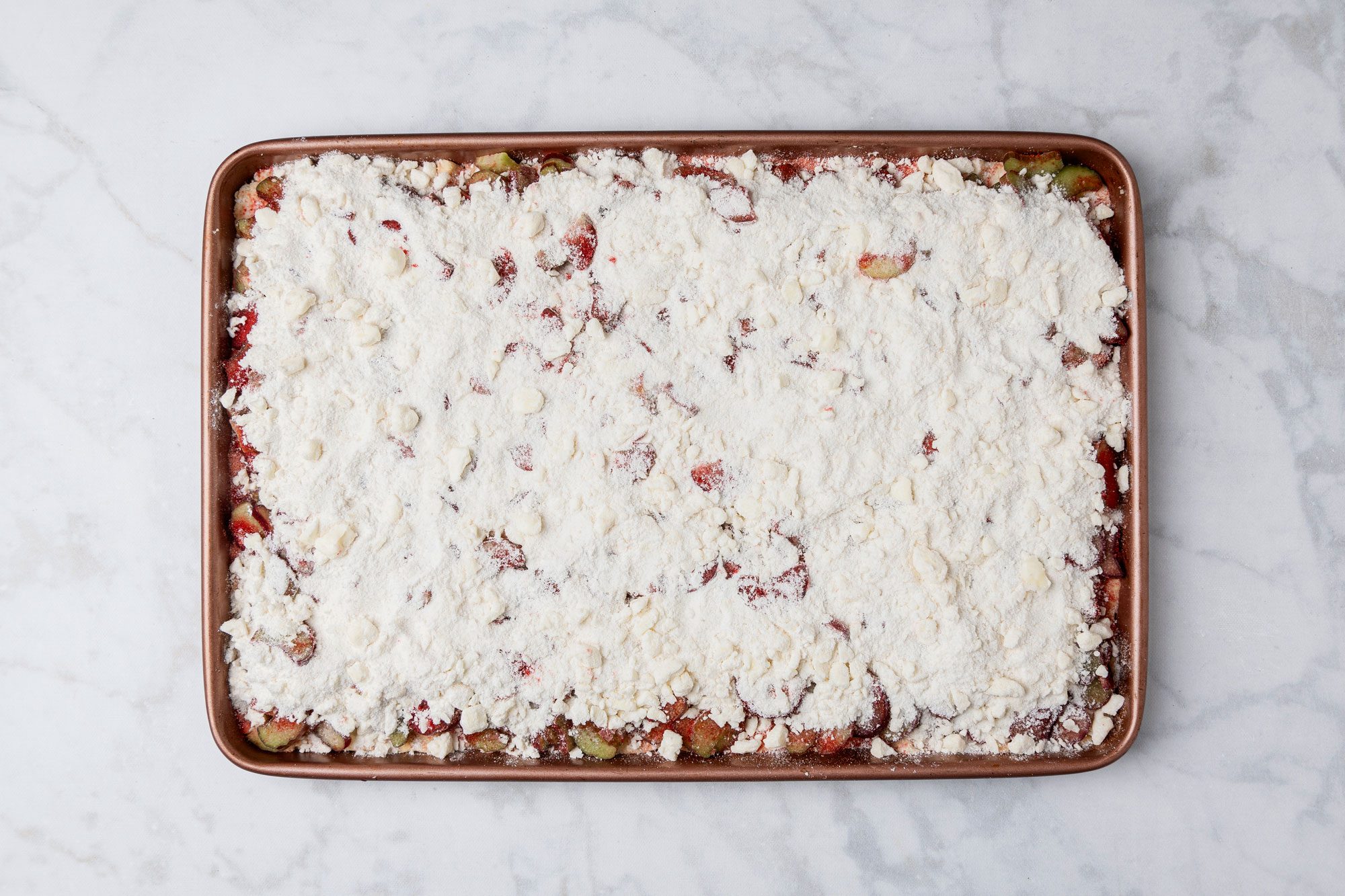 overhead shot of a rectangular baking sheet filled with an unbaked dessert, topped with a thick, crumbly layer of white flour mixture, Bits of fruit are partially visible underneath, The sheet is on a light marble surface