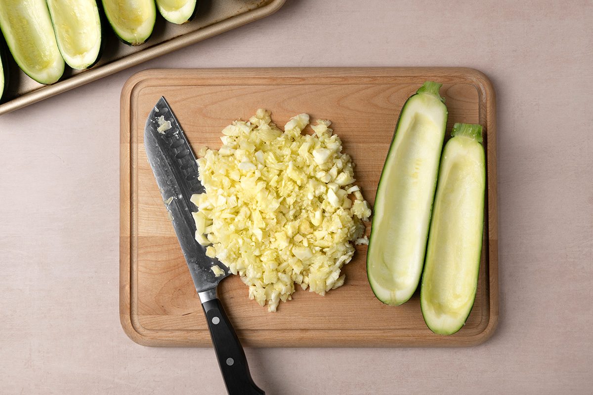 A cutting board with a knife, chopped zucchini, and two zucchini halves with their centers scooped out. A baking tray with more scooped zucchini halves is visible in the top left corner.