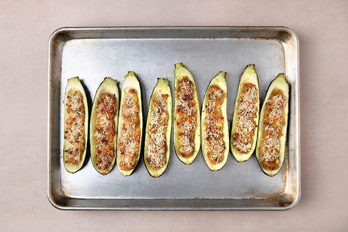 Eight zucchini halves, stuffed and topped with cheese and breadcrumbs, are arranged in a row on a baking sheet before baking.