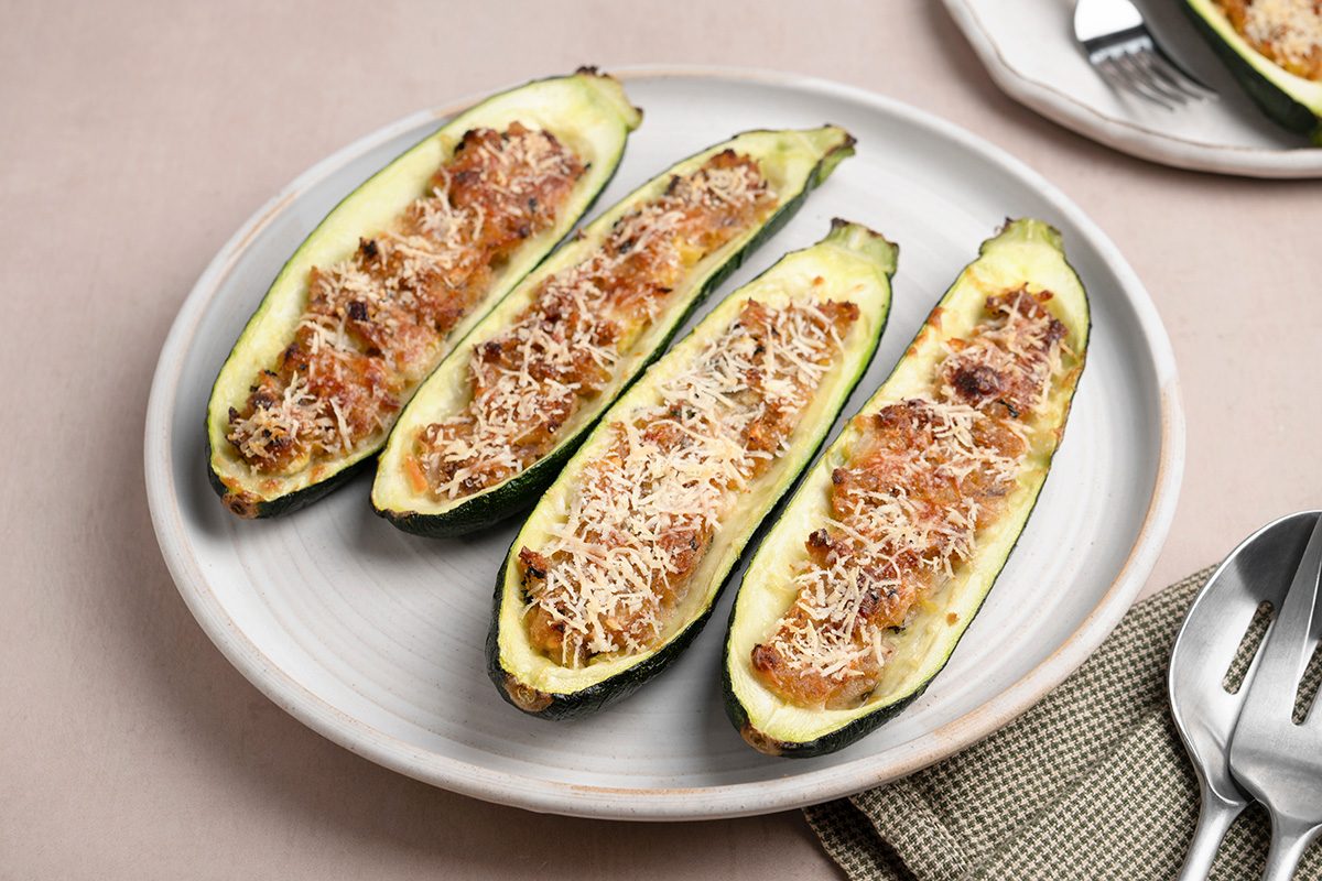 Four zucchini halves stuffed with a savory filling and topped with melted cheese are arranged on a white plate, ready to eat. A fork and spoon rest on a napkin nearby.
