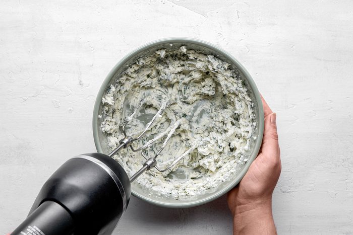 A Top Shot of a person mixes a creamy; green-flecked mixture with a hand mixer in a bowl on a light surface; suggesting food preparation