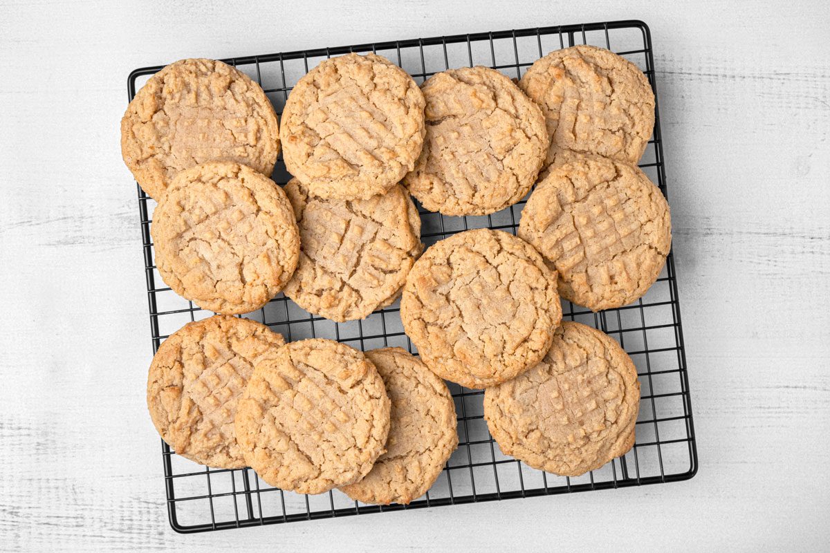 Top Shot of a cooling rack holds twelve golden-brown honey peanut butter cookies in a loose layer; set atop a white wooden surface, the background gives a bright, rustic feel