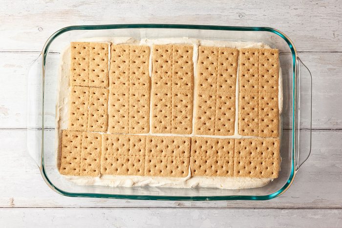 A glass baking dish filled with a layer of rectangular graham crackers placed on top of a creamy filling, sitting on a white wooden surface.