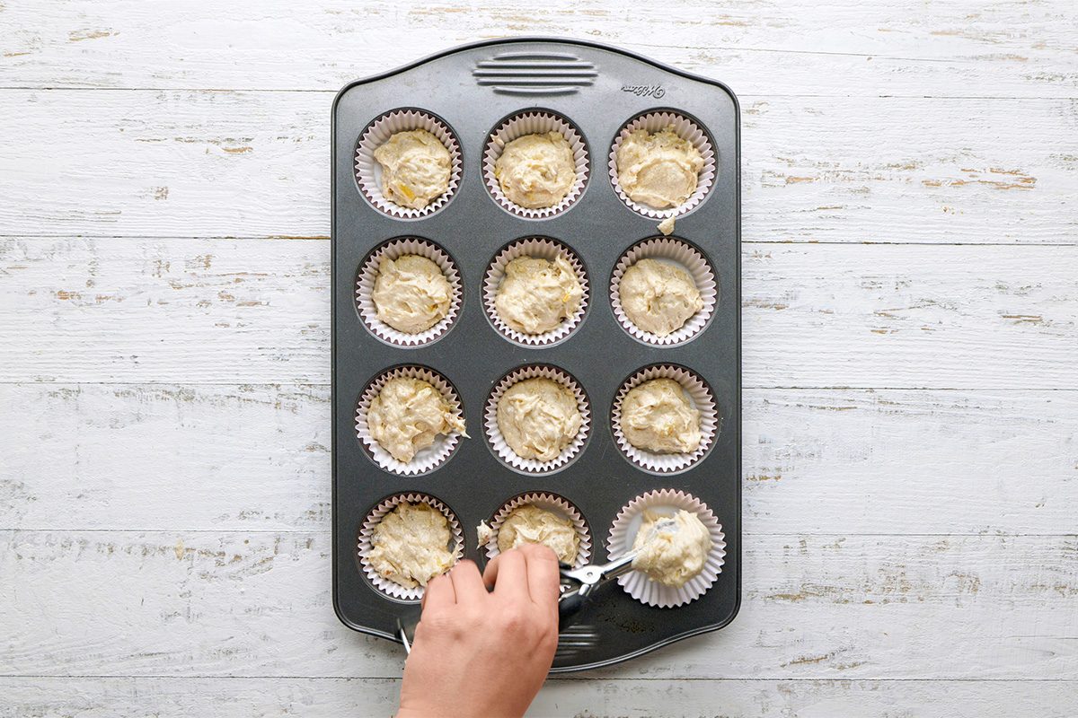 A hand uses a spoon to fill paper cupcake liners with batter in a 12-cup muffin tin, set on a white wooden surface.