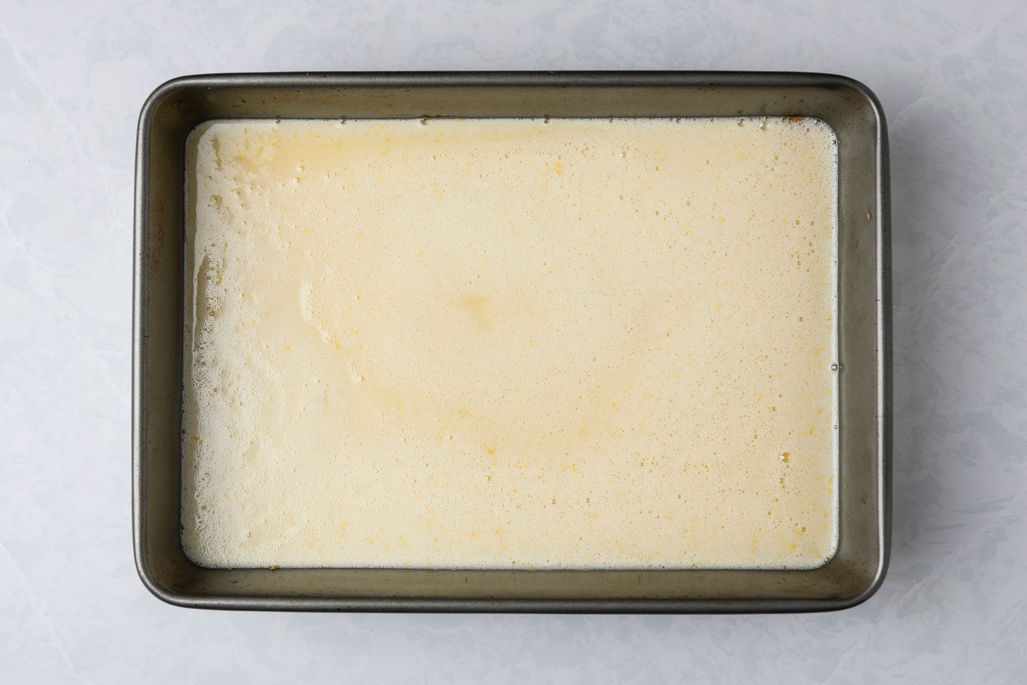 A top shot of a rectangular metal baking pan holds a pale; frothy batter; resting on a light-colored; marbled counter. The mixture looks smooth and ready for baking; suggesting preparation for cake or bread
