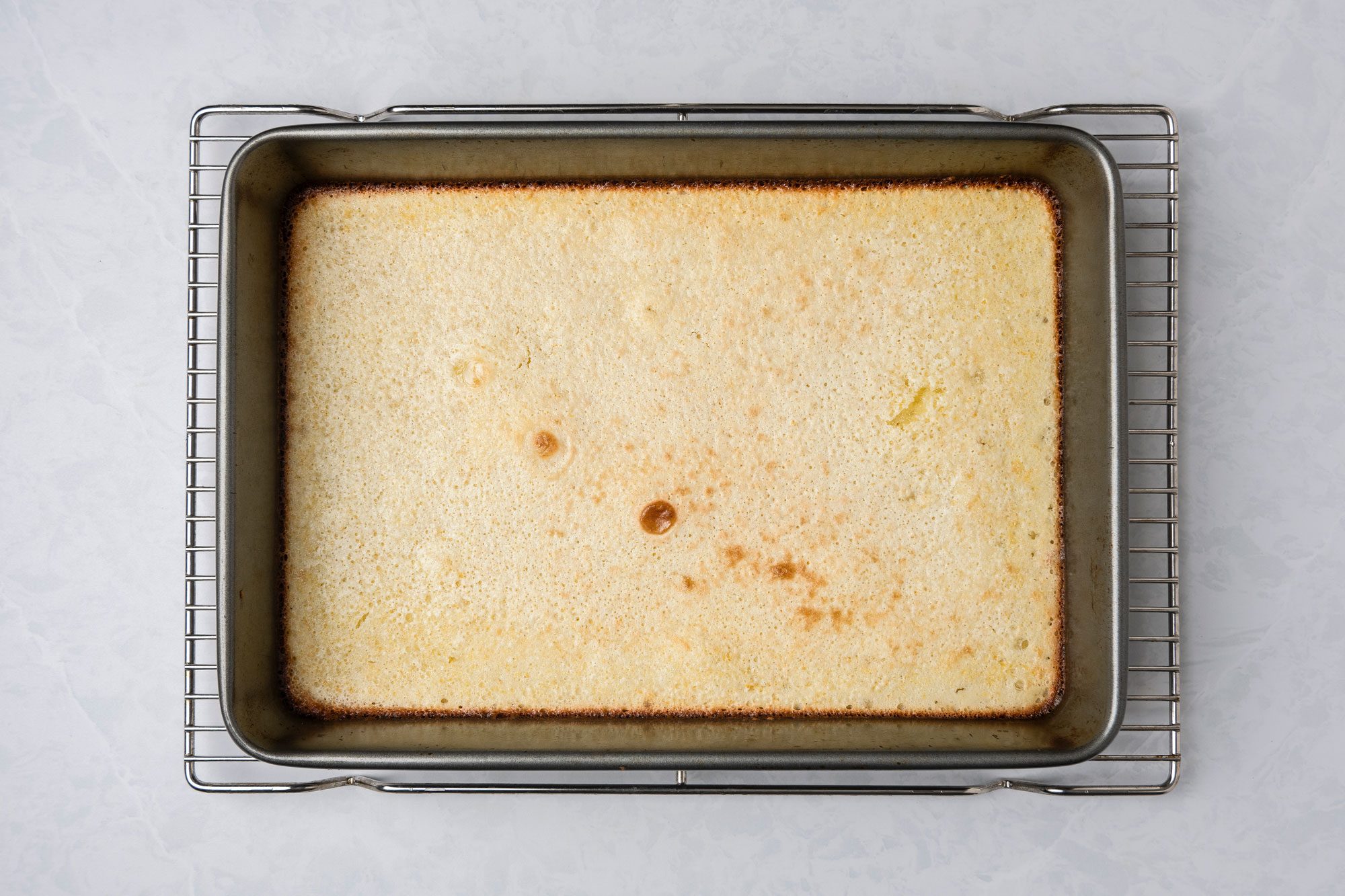 A top shot of a rectangular baking pan holds golden-brown baked custard on a cooling rack; the setup rests on a light surface, suggesting freshness and homemade comfort