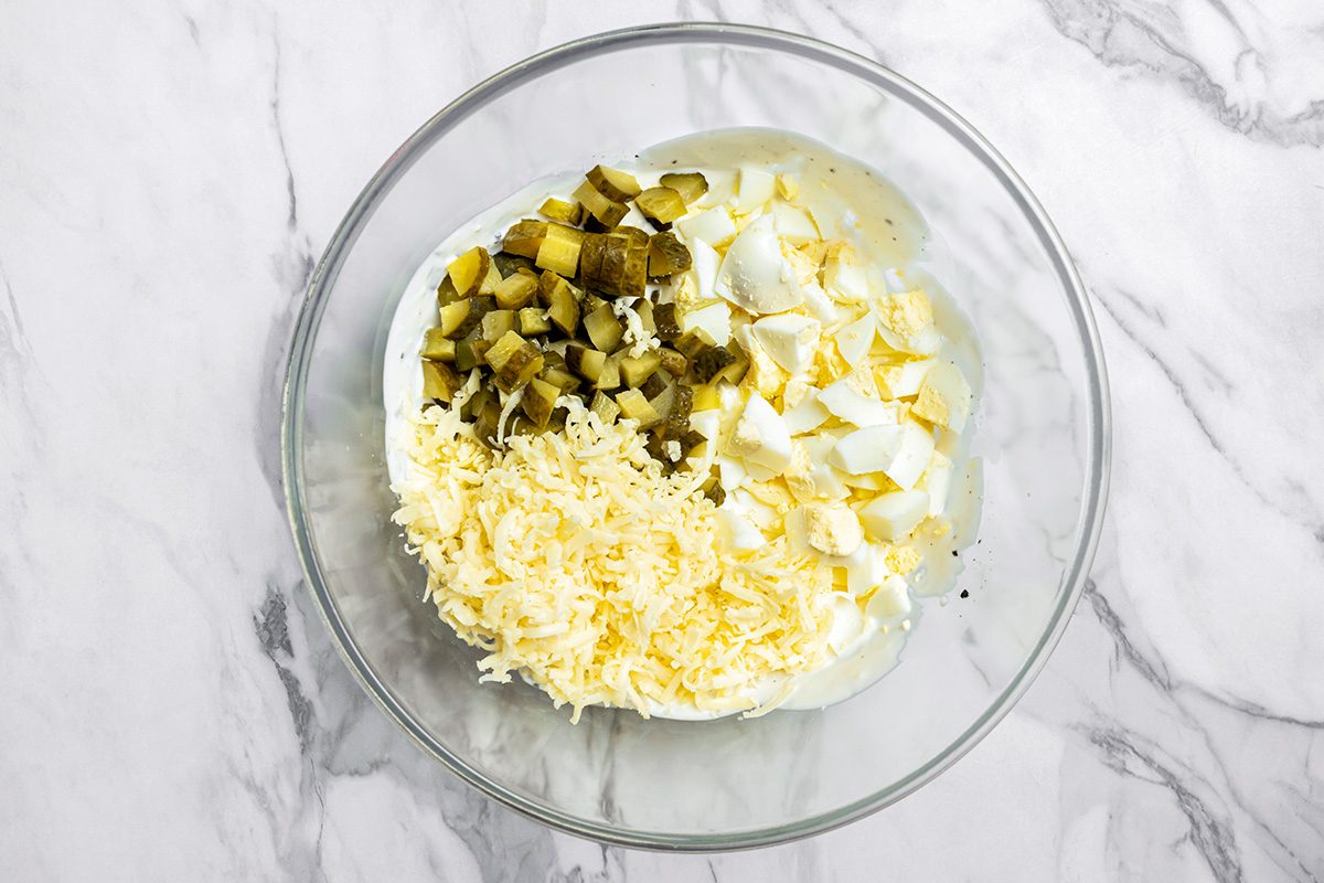 A glass bowl with chopped pickles, diced hard-boiled eggs, and grated cheese on a marble countertop, viewed from above.
