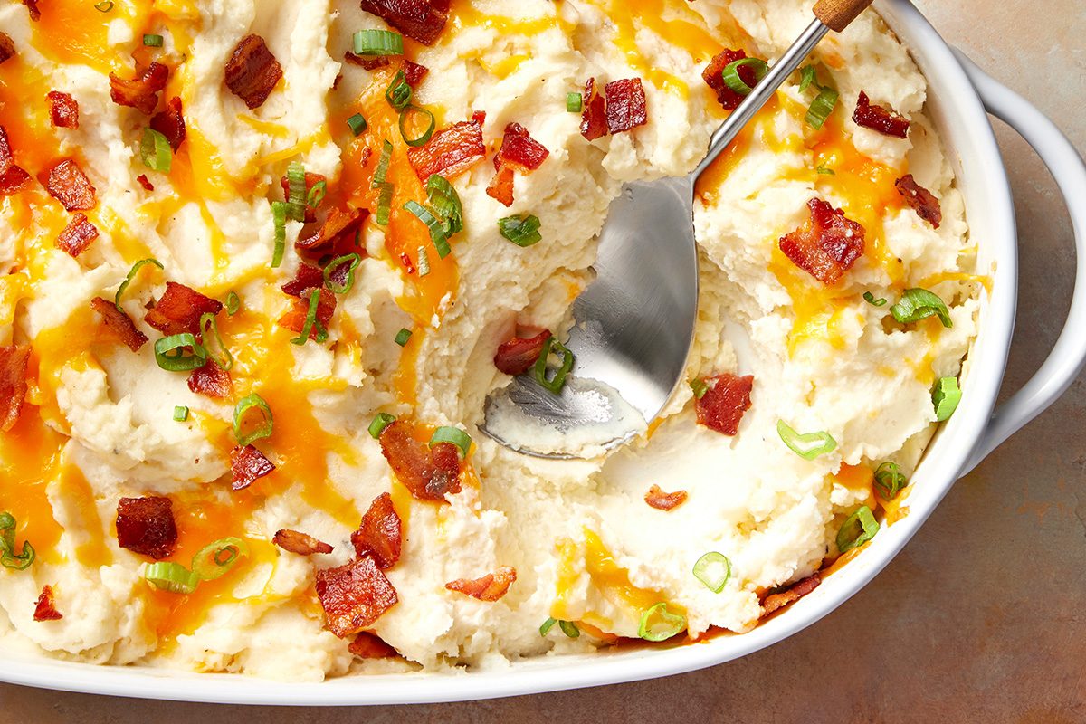 A casserole dish filled with creamy mashed potatoes topped with melted cheese, crispy bacon pieces, and chopped green onions, with a serving spoon scooping from the center.