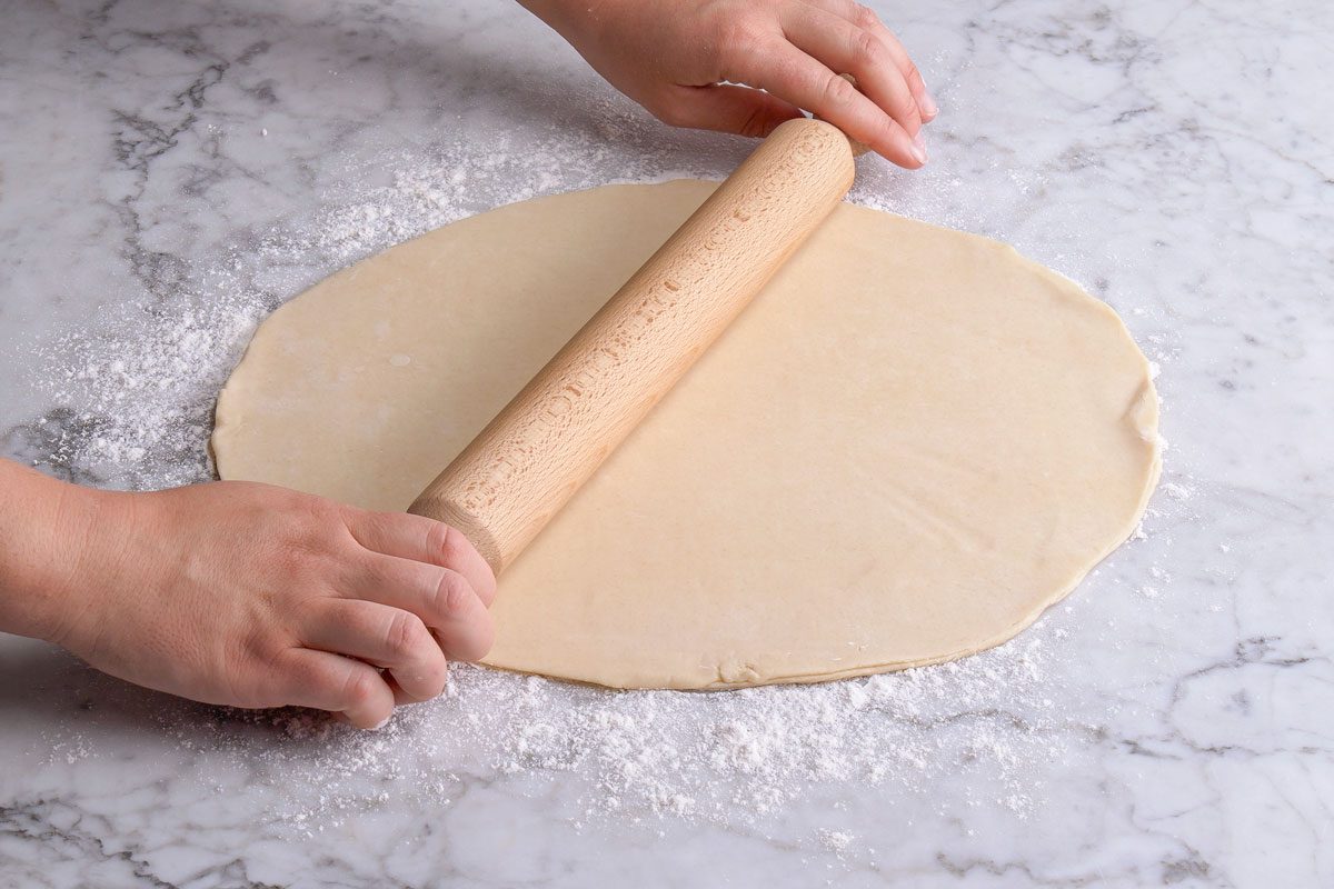 3/4th closeup shot of Hands rolling out dough with a wooden rolling pin on a floured marble surface