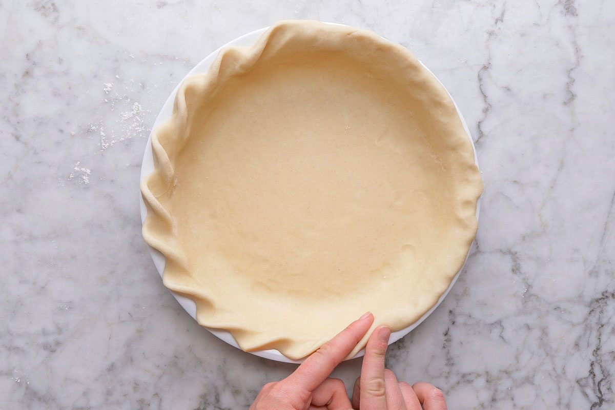 overhead shot of a person pressing the edges of an unbaked pie crust into a white pie dish on a marble countertop