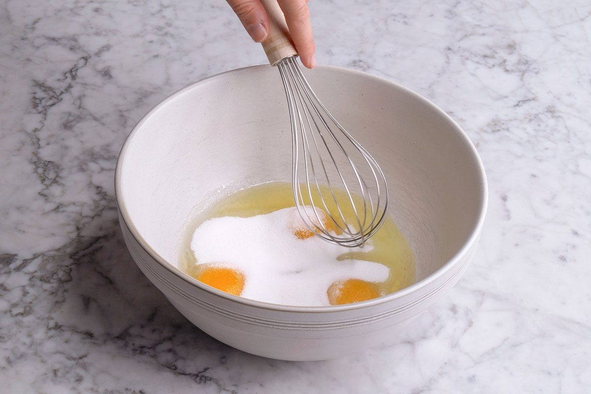 3/4th shot of a hand holds a whisk over a white bowl containing egg yolks, egg whites, and a mound of sugar on a marble countertop