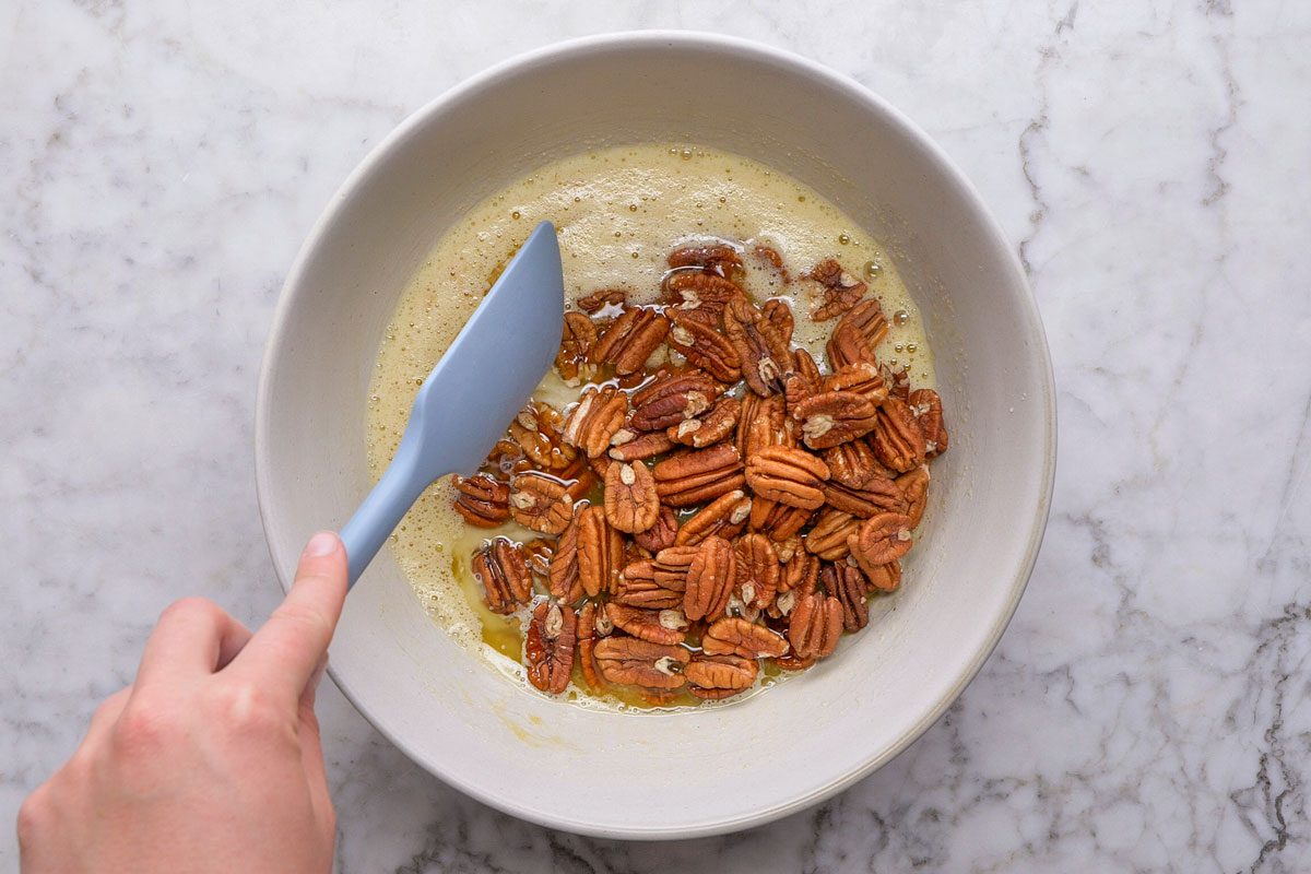 overhead shot of a hand stirs whole pecans into a bowl of frothy liquid with a light blue spatula, on a white marble countertop
