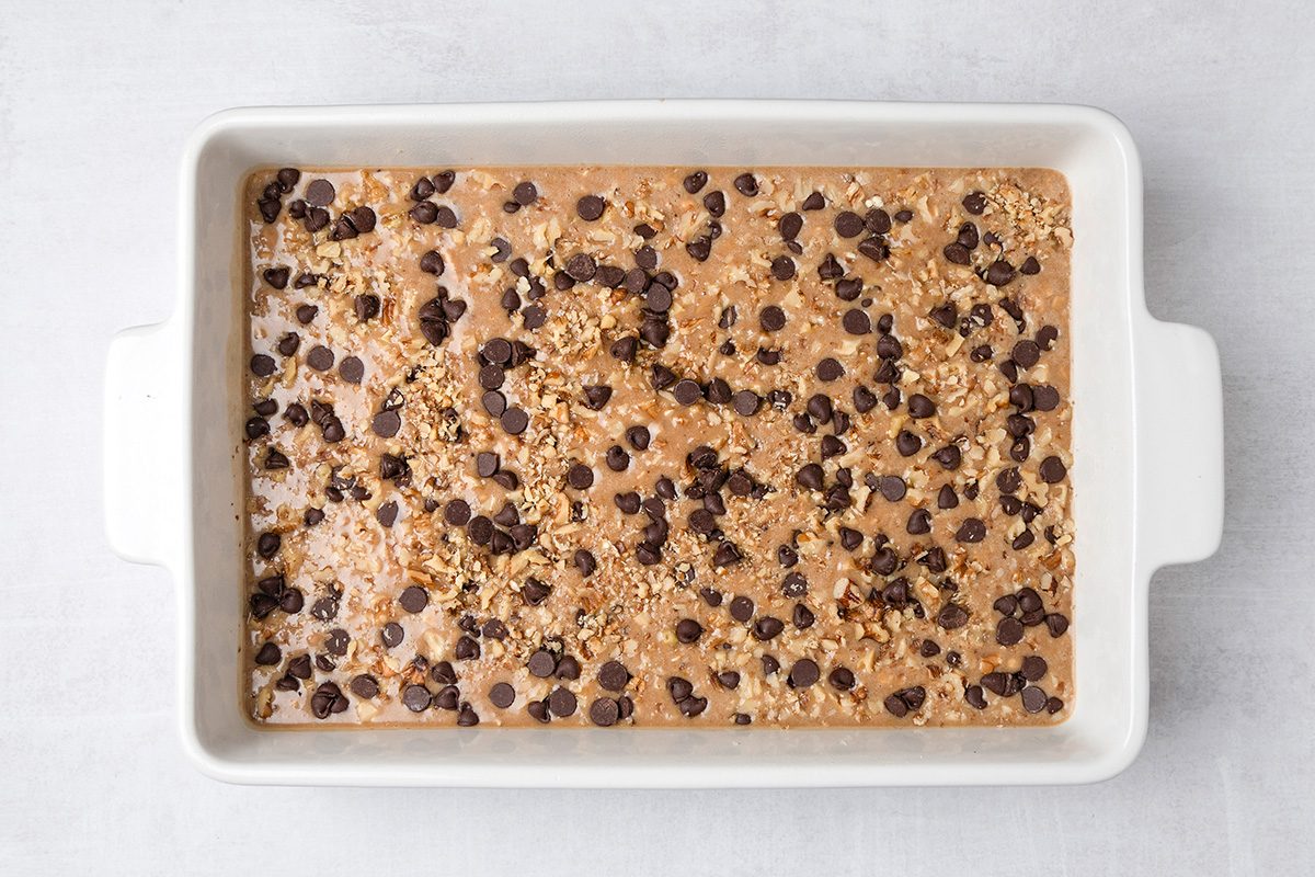 A rectangular white baking dish filled with unbaked batter, topped with chocolate chips and oats, sits on a light-colored surface.
