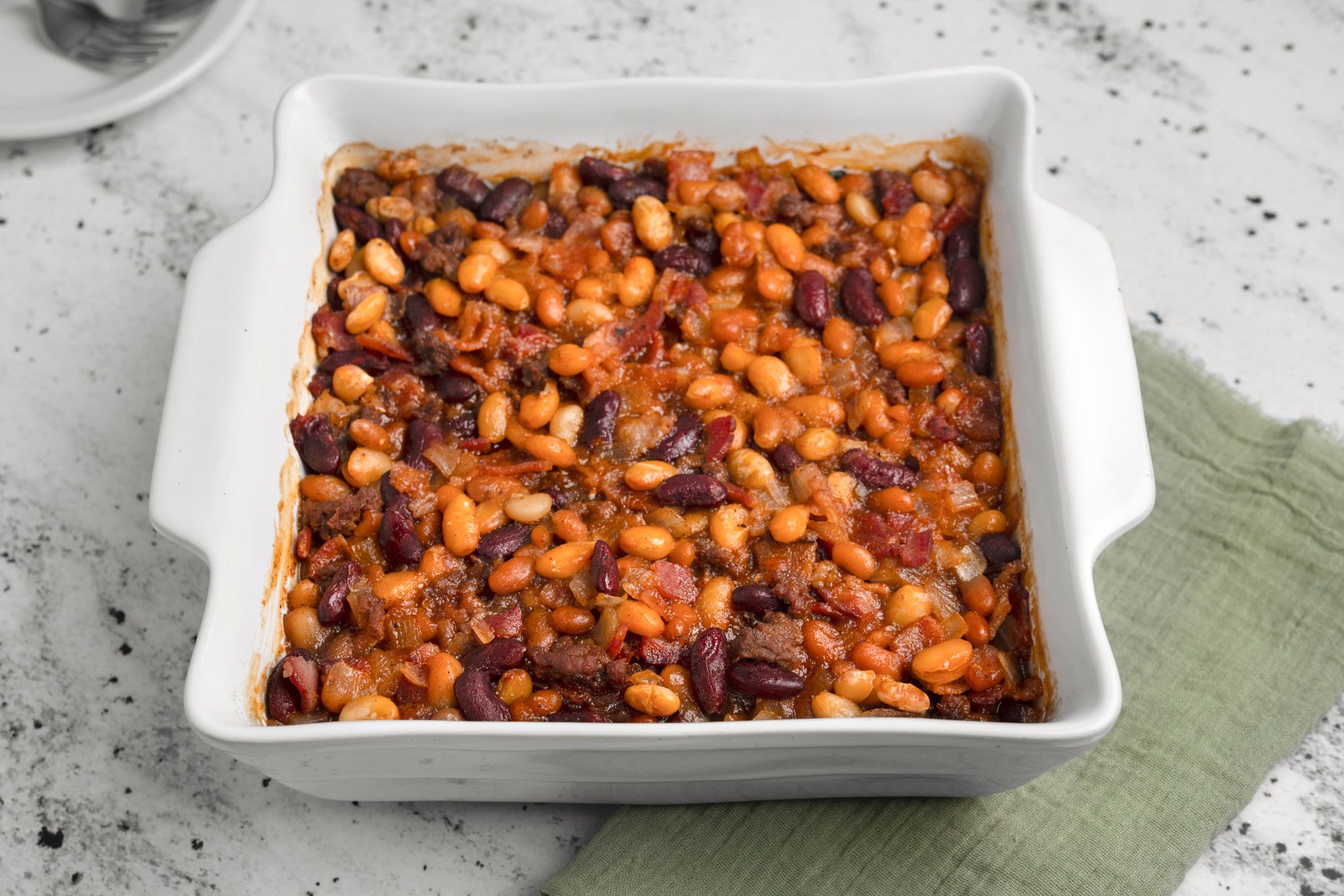 3/4th Shot of a square white baking dish contains baked beans in a tomato sauce with kidney and white beans; it rests on a light countertop beside a green cloth napkin, creating a warm, homey atmosphere