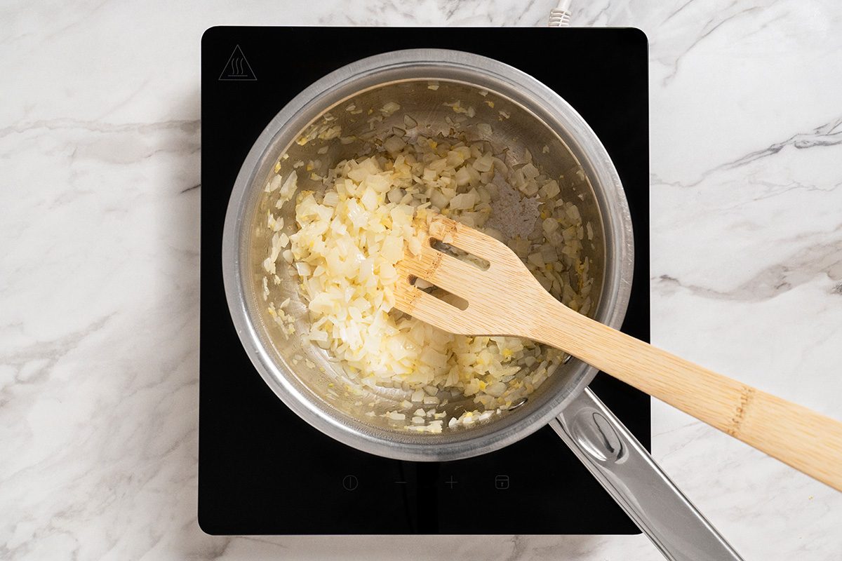 A pot with chopped onions being sautéed, stirred with a wooden spatula, sits on an electric stovetop with a marble countertop background.