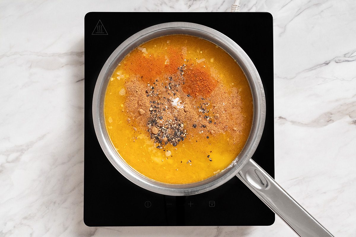 A saucepan filled with yellow soup or sauce topped with various spices sits on a black electric stovetop on a white marble surface.