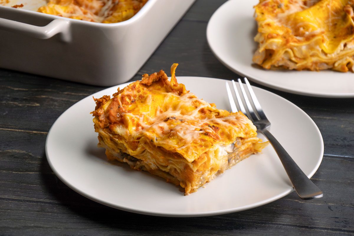 3/4 th Shot of A slice of Pumpkin Lasagna; topped with melted cheese; rests on a white plate with a fork; beside another plate and a baking dish; all arranged on a dark wooden table, creating an inviting dining scene