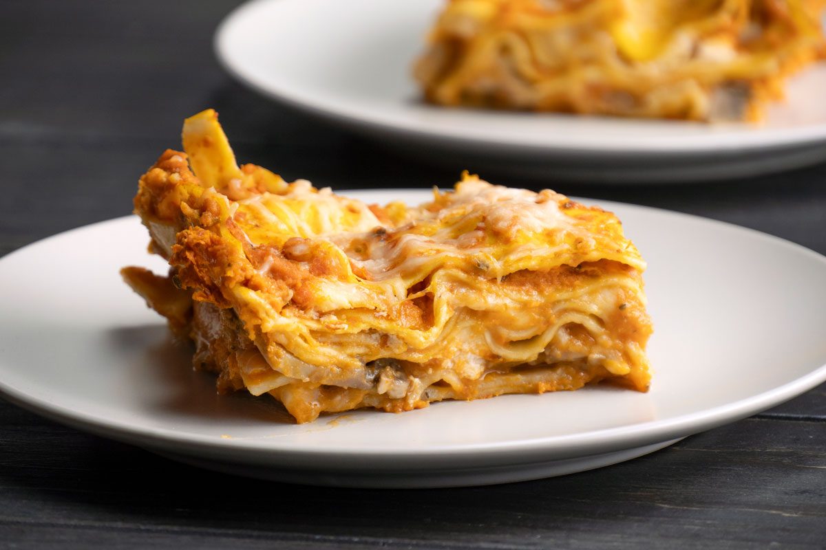 Front Shot of A close-up shows a slice of Pumpkin Lasagna featuring layers of pasta; cheese; and sauce on a white plate; with another lasagna plate softly blurred in the background