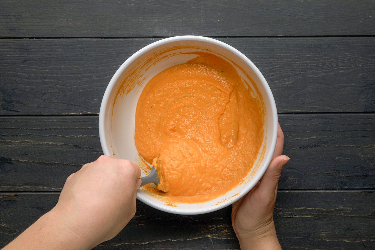 Top Shot of A person stirs vibrant orange batter in a white bowl using a spatula; the bowl rests on a dark wooden surface, creating contrast between the bright mixture and rich background tones