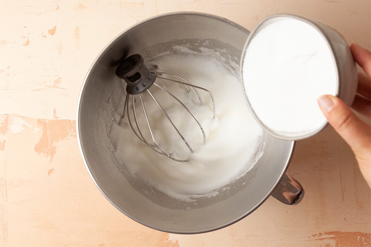 A hand holds a bowl of whipped egg whites above a mixing bowl with a whisk attachment, which already contains some whipped egg whites.